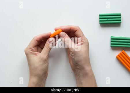 Le mani della donna che fanno l'argilla piccola del polimero della carota sullo sfondo bianco Foto Stock
