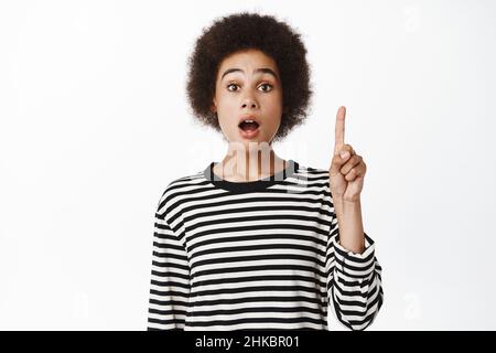 Donna nera eccitata che mostra il senso, puntando il dito in su, dicendo le notizie grandi, il movimento di eureka, levandosi in piedi sopra il fondo bianco nei vestiti casual Foto Stock