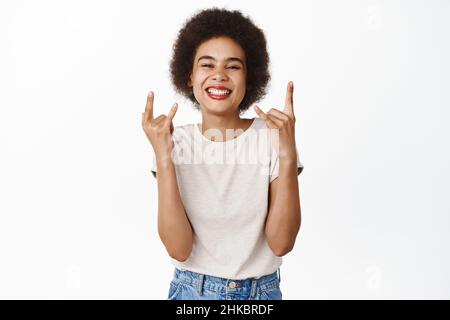 Rock on. Sorridendo ragazza africana con capelli afro, mostrando sassy segno di metallo pesante, rock sul gesto, godendo e divertirsi, in piedi su sfondo bianco Foto Stock