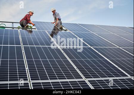 Basso angolo di due uomini tecnici solari costruire pannelli solari fotovoltaici sotto il cielo blu. Lavoratori maschi in caschi di sicurezza che collocano il modulo solare su binari metallici. Concetto di fonti di energia alternative. Foto Stock