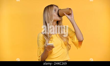 Donna bionda che beve il caffè per andare e tenere smartphone isolato sul giallo Foto Stock