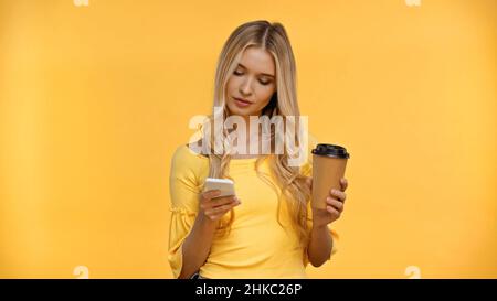 Donna bionda che usa lo smartphone e che tiene la tazza di carta isolata sul giallo Foto Stock