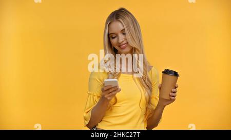 Donna bionda positiva usando lo smartphone e tenendo il caffè per andare isolato sul giallo Foto Stock