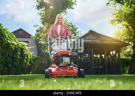 Vista ad angolo basso di donna caucasica sorridente con capelli biondi usando il rasaerba elettrico mentre lavora in giardino. Attraente donna in abiti casual taglio erba con attrezzature da giardinaggio moderno. Foto Stock