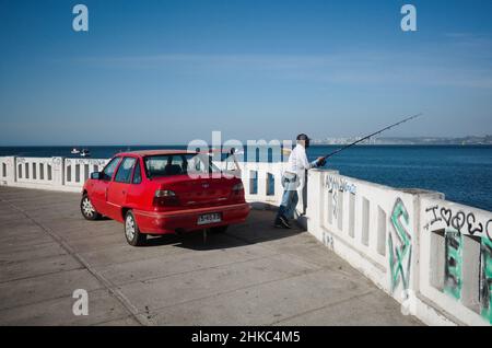 Valparaiso, Cile - Febbraio, 2020: L'uomo con canna da pesca è la pesca sul lungomare della città di Valparaiso. L'auto Red Daewoo è parcheggiata su un argine Foto Stock