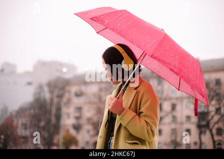 Giovane donna premurosa in cuffie con ombrello che cammina in città Foto Stock