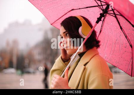 Giovane donna premurosa in cuffie con ombrello che cammina in città Foto Stock