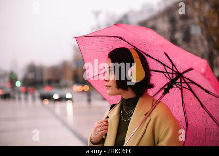 Giovane donna premurosa con cuffie che tengono l'ombrello in piedi in città Foto Stock