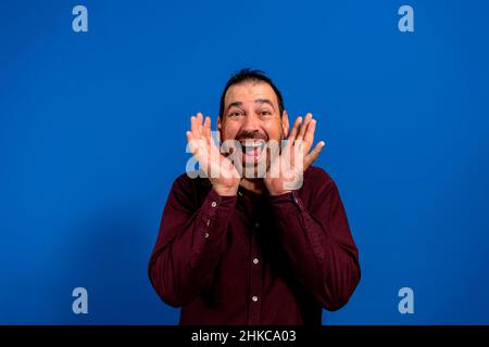 Giovane bell'uomo in camicia viola su sfondo blu sorridendo felicemente giocando un boo con le mani che mostrano il volto. Sorpreso e. Foto Stock