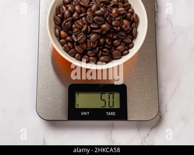 Chicchi di caffè arrosto in una ciotola su bilancia digitale da cucina. La bilancia visualizza 50g di chicchi di caffè senza tara. Dispositivo elettronico metallico. Foto Stock