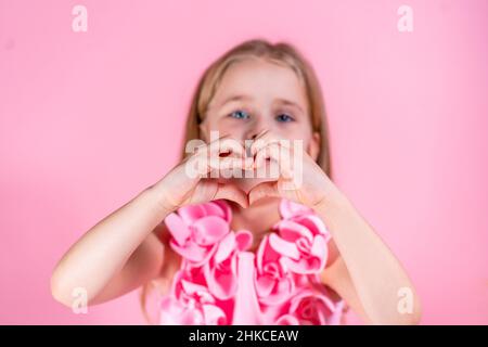 Ritratto di bella ragazza con occhi blu con capelli lunghi e leali in elegante vestito rosa posa che mostra la forma del cuore con le mani. Foto Stock