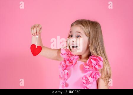 Ragazza carina con occhi blu e lunghi capelli lisci in elegante vestito rosa stand sorridente guardando rosso cuore di carta sul filo. Foto Stock