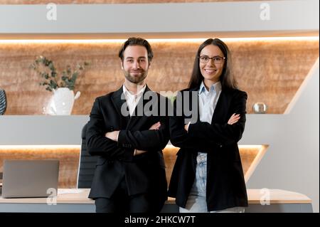 Successo intelligente caucasico giovane uomo e donna adulti, colleghi di ufficio, finanza, imprenditori, in piedi al desktop, in abiti formali ed eleganti, braccia incrociate, amichevole sorridente Foto Stock