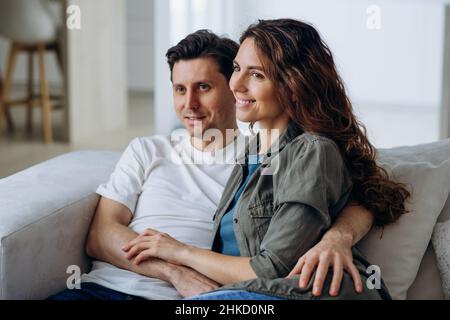 Sposi felici con capelli scuri sedersi su un comodo divano in soggiorno e discutere i piani per il futuro sorridendo ampiamente primo piano Foto Stock