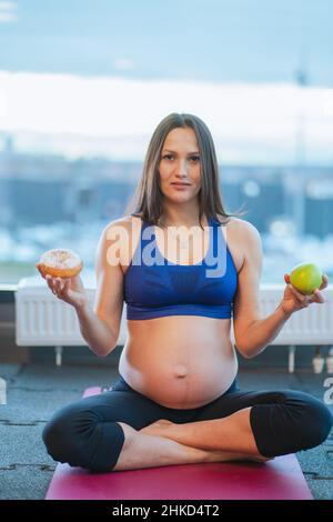 Giovane donna incinta beauteous in abbigliamento sportivo blu si siede su tappeto rosa scegliendo tra mela e ciambella in palestra. Verticale. Foto Stock