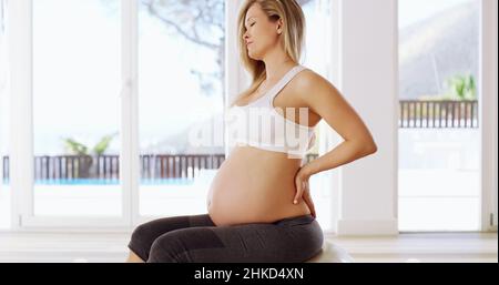 Darle un po' di riposo. Scatto corto di una giovane donna incinta attraente seduta con le mani sulla schiena. Foto Stock