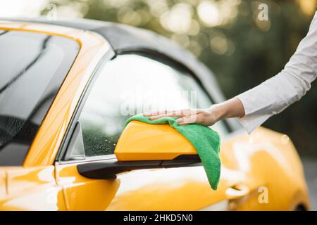 Raccolto primo piano di mano di giovane donna caucasica tergendo specchietto retrovisore auto della sua moderna auto di lusso in una stazione di lavaggio auto self-service all'aperto con un panno in microfibra verde in estate mattina soleggiato Foto Stock