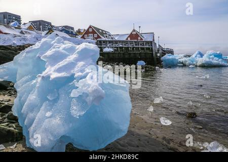 Grande pezzo di ghiaccio blu che stabilisce tra i sassi sulla riva con Inuit moderno edificio sulla collina al fiordo, città di Nuuk, Groenlandia Foto Stock