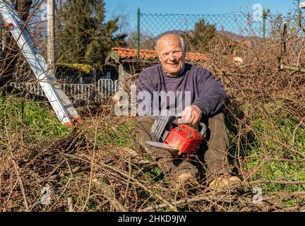 Uomo anziano felice con motosega seduta a riposo dopo aver tagliato l'albero in giardino Foto Stock