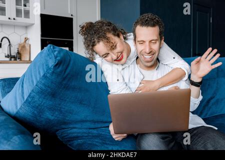 Coppia di matrimonio gioiosa che hanno una videochiamata da parenti o amici durante la quarantena, sorridendo. L'uomo felice seduto sul divano e salutando online Foto Stock