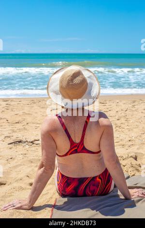 Donna matura in un costume da bagno si siede su una spiaggia sabbiosa e guarda il mare, vista posteriore. Resto dei pensionati. Foto Stock