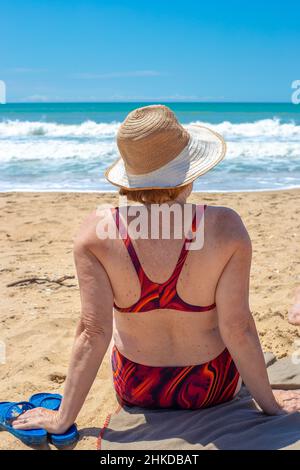 Donna matura in un costume da bagno si siede su una spiaggia sabbiosa e guarda il mare, vista posteriore. Resto dei pensionati in Russia. Foto Stock