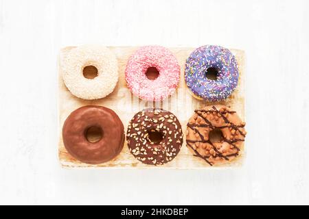 Sei ciambelle dolci smaltate colorate su un asse di legno bianco. Vista dall'alto, spazio di copia per il testo Foto Stock