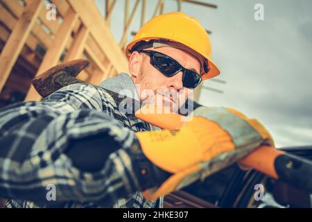 Caucasico professionista Construction Contractor Worker nel suo 40s con un martello grande sulla spalla. Indossare cappello rigido giallo e protezione occhi Sungla Foto Stock