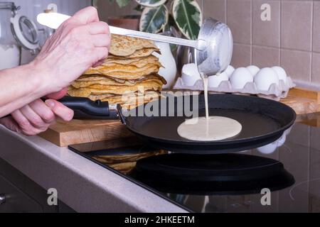 La donna casalinga cuoce le frittelle per la colazione e versa l'impasto in una padella calda. Pancakeson fatti in casa in vetroceramica stufa in cucina. Ingredienti per Foto Stock