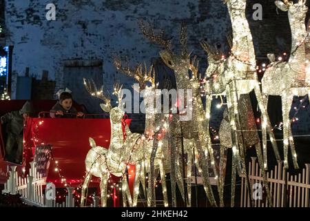 Prattville, Alabama, USA- 23 dicembre 2021: La bambina si siede in slitta rossa con suo fratello e guarda la renna illuminata in stupore nella storica d. Foto Stock