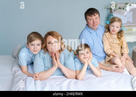 Famiglia felice che guarda la macchina fotografica sul loro letto in camera da letto. I genitori e i bambini caucasici si mentono e si divertono a casa nel loro tempo libero. Grande famiglia amichevole stanno riposando in vacanza. Figli e dolce figlia Foto Stock