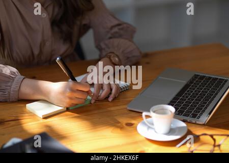 Scatto ritagliato della studentessa dell'università femminile che prende nota sul taccuino bianco mentre cerca le informazioni sul laptop nella biblioteca o nel coffee shop. Foto Stock