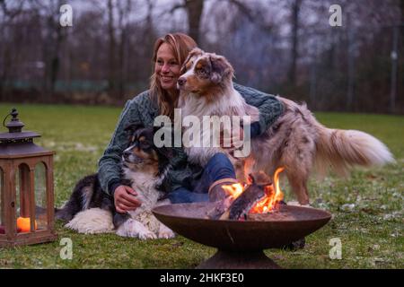 La giovane donna è seduta fuori nei boschi con i suoi due cani pastorelli australiani. Neve sull'erba, crepuscolo vicino al fuoco. Foto Stock