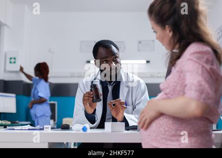 Medic afroamericano che dà una bottiglia di pillole a persona incinta per curare la malattia. Pallone di tenuta del medico con medicinale da prescrizione e trattamento da somministrare a una donna in gravidanza. Foto Stock