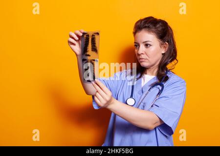 Infermiere donna che guarda la radiografia per trovare la diagnosi sanitaria. Assistente medico che indossa una protezione e uno stetoscopio uniformi durante la scansione radiografica e l'analisi dei risultati Foto Stock