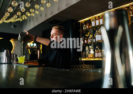 Vista laterale ad angolo basso del barman che versa una bevanda alcolica fresca in bicchieri con cubetti di ghiaccio dietro il bancone del bar nel moderno nightclub. Foto Stock