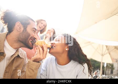 Divertente foto di una coppia che mangia un croissant come scherzo mentre gli amici guardano sullo sfondo. Foto Stock