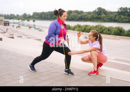 Ampio scatto di obese in sovrappeso giovane donna facendo esercizi utilizzando nastro fitness per la perdita di peso con personal trainer all'aperto nella giornata estiva. Foto Stock