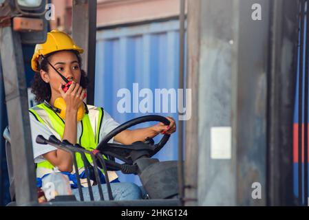 Il caposquadra femmina usa la comunicazione radio per comunicare mentre guida il carrello elevatore al deposito del contenitore di spedizione. Spedizione in banchine. Foto Stock