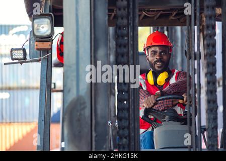 Lavoratore di sesso maschile in uniforme e casco che guida e funziona su carrello elevatore a forche con container diesel presso la banchina commerciale. Foto Stock