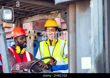 Due lavoratori afroamericani maschi e femmine in uniforme e casco che guidano e operano su carrelli elevatori a forche con container diesel presso la banchina commerciale. Foto Stock
