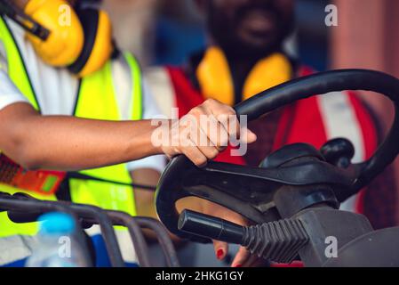 Due lavoratori afroamericani maschi e femmine in uniforme e casco che guidano e operano su carrelli elevatori a forche con container diesel presso la banchina commerciale. Foto Stock