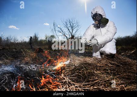 Persona irriconoscibile che brucia vecchia erba secca in campo. Fuoco su un fuoco. Ecologista che tiene il matchbox e fuoco di asciugare l'erba sotto il cielo blu, indossando tuta protettiva e respiratore. Foto Stock