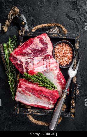 Costolette di manzo organico crudo pronte per essere cuote in vassoio di legno con erbe. Sfondo nero. Vista dall'alto Foto Stock