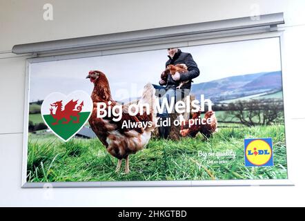 Lidl Supermarket campagna poster logo cartello con Farmer, Hen e Welsh Dragon pubblicità fuori negozio nella primavera Febbraio 2022 Galles Regno Unito Gran Bretagna Foto Stock