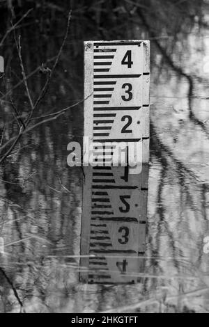 Misuratore di profondità dell'acqua in piccolo bianco fiume con letture in nero parte sommersa conversione in bianco e nero in bianco e nero in primo piano ramoscelli sullo sfondo Foto Stock