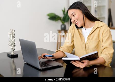 Multitasking. La donna freelance asiatica parla sullo smartphone e prende appunti con la penna, utilizzando un computer portatile per il lavoro remoto da casa, scrivere alcune informazioni seduti al tavolo Foto Stock