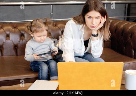 Seria giovane donna d'affari in maternità lasciare seduta sul divano con il bambino piccolo, utilizzando il portatile, facendo rapporti importanti. Bella madre che lavora da Foto Stock