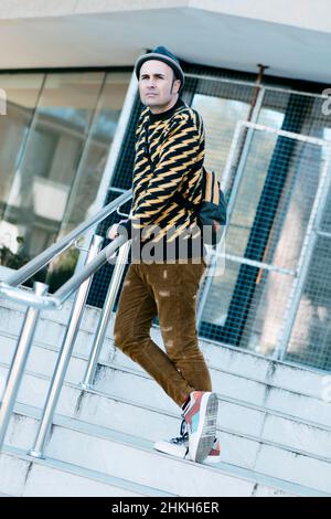 uomo di mezza età, con un paio di moderni pantaloni corduroy marroni, un pullover a motivi geometrici marrone e nero e un cappello, in piedi su una scala esterna Foto Stock