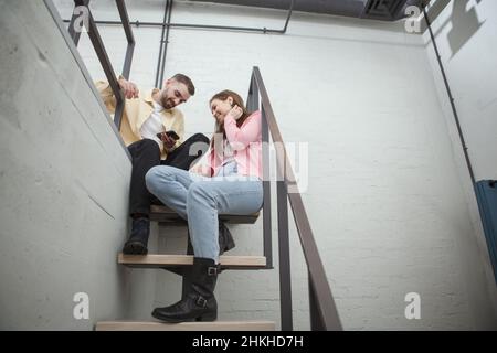 Scatto ad angolo basso di una bella giovane coupke chiacchierando, usando lo smart phone seduto in cima alle scale nella camera d'albergo, spazio copia Foto Stock